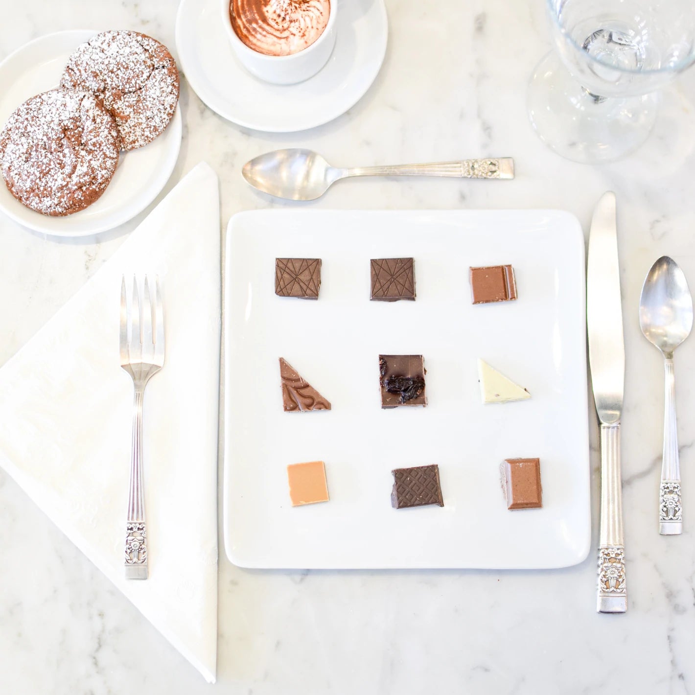A set table with a variety of chocolate pieces on a white plate, accompanied by a cup of coffee and pastries, arranged for a tasting event.